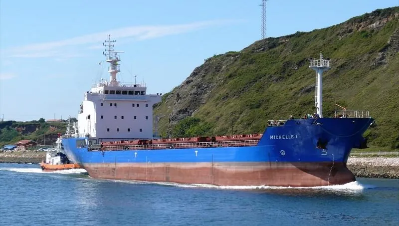 Michelle 1 under way along a hilly coastline, escorted by a tug