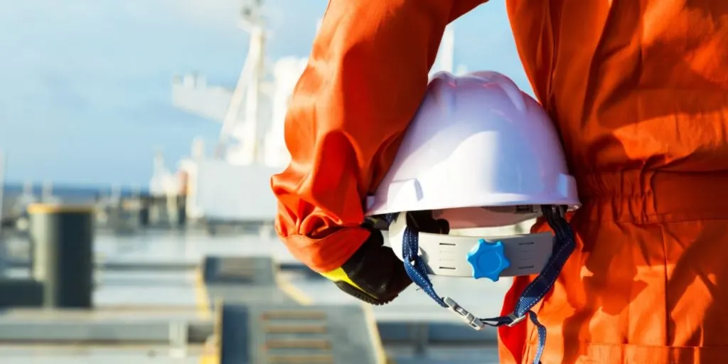 A seafarer in orange coveralls holding a white safety helmet